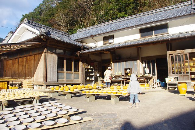 This is an Onta Ware workshop in the village of Onta, Oita, Japan. In the front yard of the workshop, a lot of formed bowls are drying in the sun on wooden boards. Families are visiting the workshop.