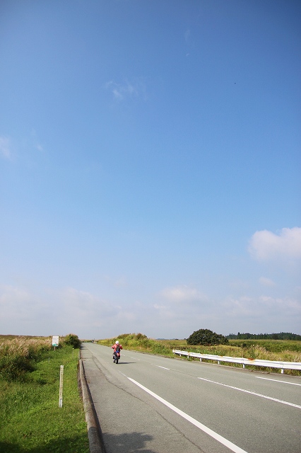 The Yamanami Highway is a road that connects Yufuin in Oita to Aso in Kumamoto in Japan. Under the blue sky, a motorcycle is riding along a straight road in the grassland.