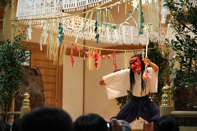 This is the Takachiho Kagura (Totori-no-mai dance) in the Kaguraden Hall on the grounds of Takachiho Shrine in Miyazaki, Japan. Tajikarao no mikoto, wearing a red mask and hakama, is holding a staff and performing a powerful dance.