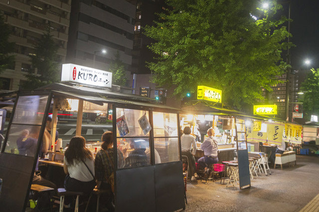 At night, there are several food stalls lined up on the sidewalk in Fukuoka, Japan. Customers inside the stalls are enjoying eating and drinking.